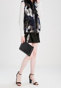 Frau trägt eine gemusterte Pelzweste, weiße Bluse, schwarzen Leder-Minirock, hält eine schwarze Clutch mit Nieten und trägt schwarze Sandalen mit hohen Absätzen.