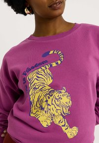 Mujer vistiendo un sudadera púrpura con un gráfico de tigre en amarillo y azul marino y el texto "el amor es libertad" en la parte frontal, con aros de oro visibles.