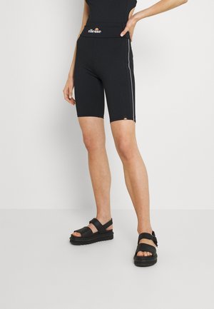 Femme portant un short noir taille haute avec des bandes blanches latérales et des sandales noires, debout avec une main sur la hanche sur un fond uni.
