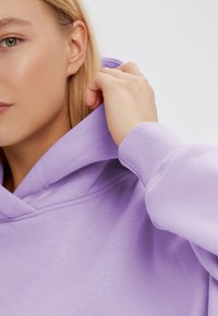 Felpa viola chiaro con cappuccio regolabile, polsini a coste e texture morbida. Mano che afferra il cappuccio, evidenziando il design rilassato.