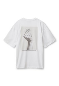 Camiseta de algodón blanca con un corte relajado. Presenta un gráfico de una mano haciendo un gesto de corazón en la parte posterior, acompañado de un texto debajo.