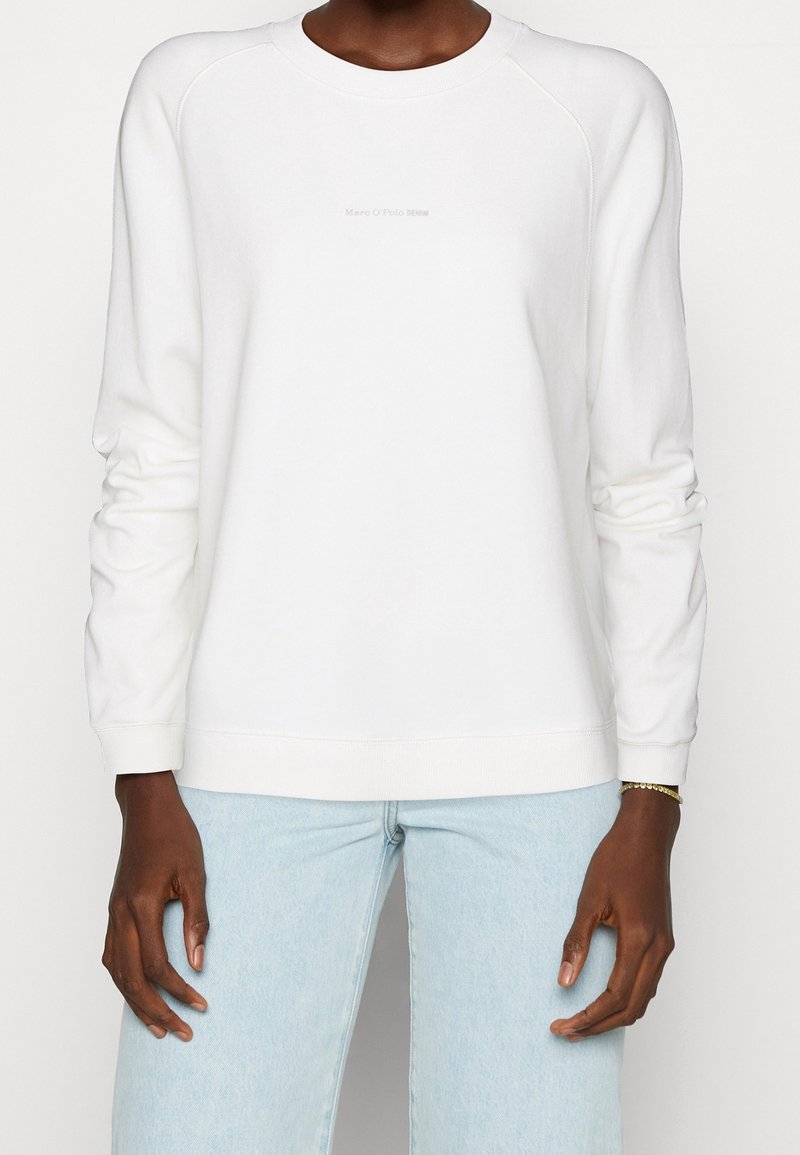 Sweat-shirt blanc avec manches raglan, col rond et logo frontal discret. Tissu doux, coupe décontractée, associé à un jean bleu clair.