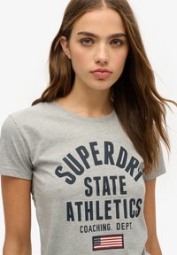 Grå T-shirt med korte ærmer, der har fed navy tekst, der står "SUPERDRY STATE ATHLETICS", samt et lille amerikansk flag grafik.