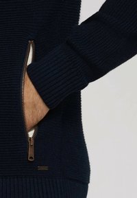 Pull en tricot bleu marine avec texture côtelée, poche zippée avec quincaillerie en bronze, et manches ajustées avec poignets côtelés.