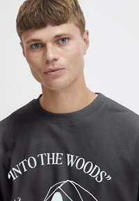 Sudadera gris con estampado gráfico que dice "EN EL BOSQUE". Presenta cuello redondo y puños de canalé. Fabricada en un tejido suave y texturizado que muestra un ligero brillo.