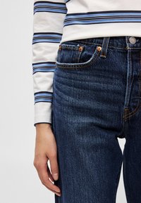 Μπλε και λευκό ριγέ μακρυμάνικο πουκάμισο συνδυασμένο με σκούρο μπλε τζιν, με πορτοκαλί ραφές και χάλκινα καρφιά στις τσέπες.