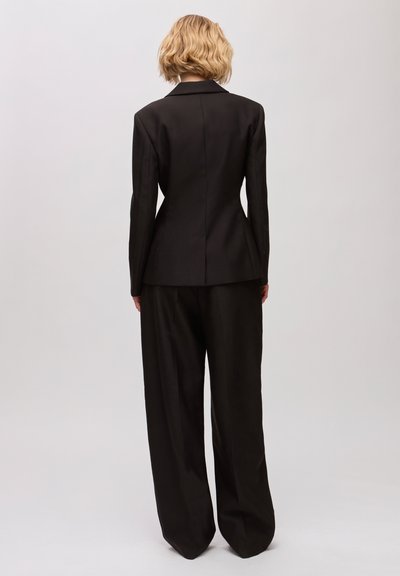 Femme aux cheveux blonds courts portant un blazer noir ajusté et un pantalon noir à jambes larges, debout de dos sur un fond clair uni.