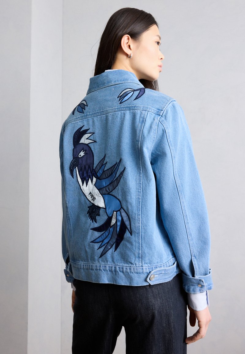 Γυναίκα που φοράει ένα ανοιχτό μπλε τζιν μπουφάν με μεγάλη κεντημένη εικόνα μπλε και λευκού πουλιού στην πλάτη, στέκεται μπροστά σε έναν απλό τοίχο.