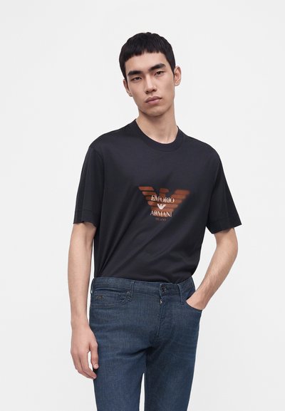 Νέος άνδρας που φοράει μαύρο t-shirt Emporio Armani με λογότυπο, σκούρο μπλε τζιν, στέκεται με το ένα χέρι στην τσέπη μπροστά σε λευκό φόντο.