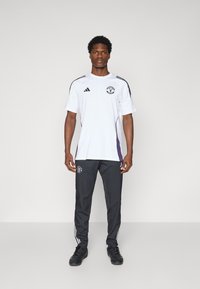 Witte sportt-shirt met zwarte en paarse accenten, met een logo op de borst. Gecombineerd met donkergrijze broek en zwarte sportschoenen.