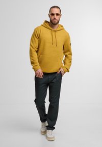 Žlutá texturovaná mikina s geometrickým vzorem, spárovaná s tmavými kalhotami a světlými teniskami. Design zahrnuje kapuci a šňůrky na stažení.