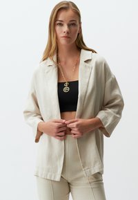 Beige Leinenblazer mit lockerer Passform, 3/4-Ärmeln und offenem Frontdesign. Kombiniert mit einem schwarzen Crop-Top und beigen Hosen. Goldene Halskette sichtbar.