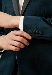 Tengerészkék bársony blazer fekete selyem hajtókával, egyetlen zsebbel és négy fekete szövetgombbal az ujjon. Fehér ing látható alatta.