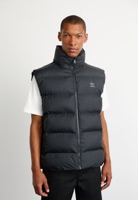 Sort puffervest med høj krave og lynlås. Har polstrede sektioner og et broderet logo på venstre bryst. Bæres over en hvid skjorte.