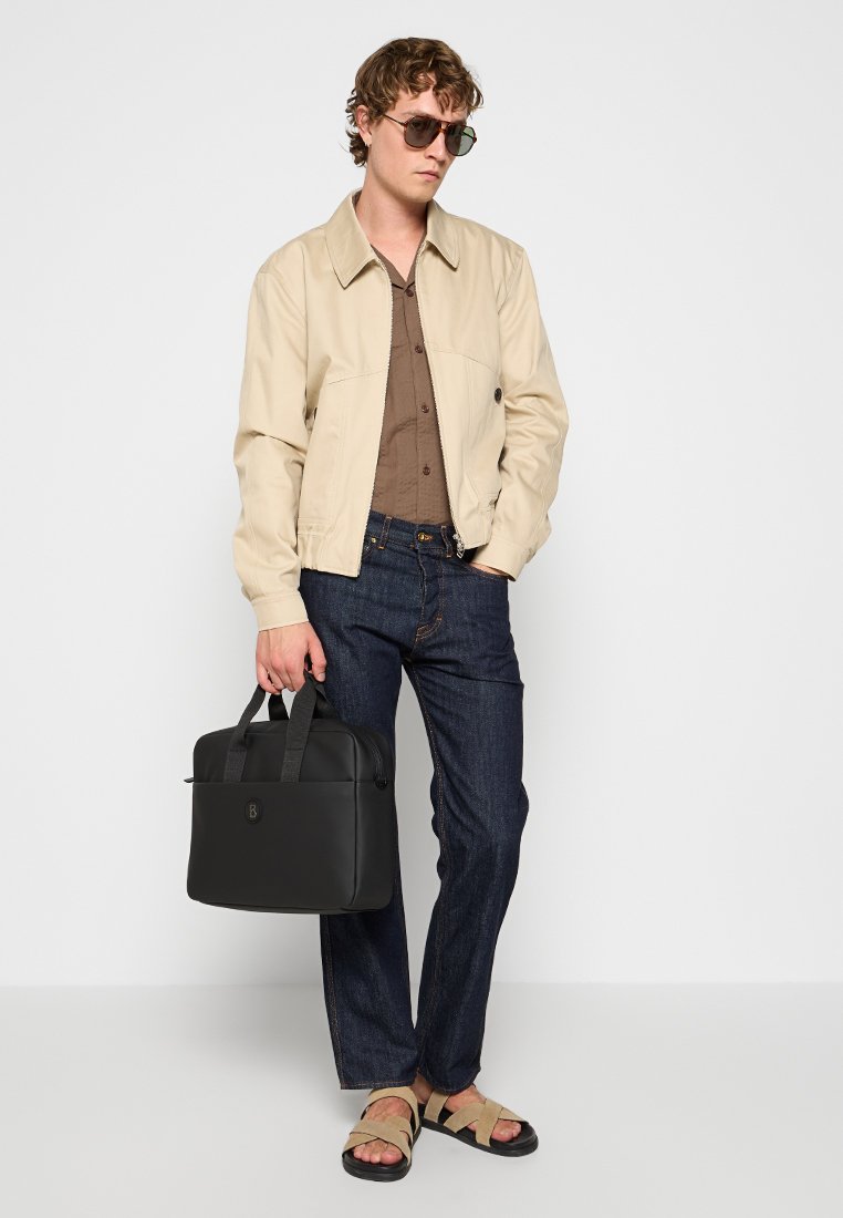 Mann trägt eine beige Jacke, ein braunes Hemd, dunkle Jeans, Sandalen und Sonnenbrille, während er eine schwarze Aktentasche vor einem weißen Hintergrund hält.