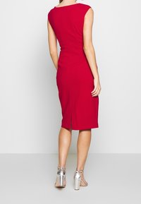 Robe rouge ajustée avec une finition au niveau des genoux, des manches courtes et une fermeture éclair dans le dos. Tissu texturé et fente à l'arrière pour le mouvement. Talons argentés visibles.