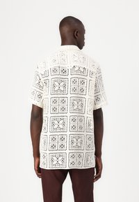 Chemise à manches courtes blanche avec des motifs géométriques découpés en noir. Silhouette décontractée avec un col, présentant des détails de couture visibles.