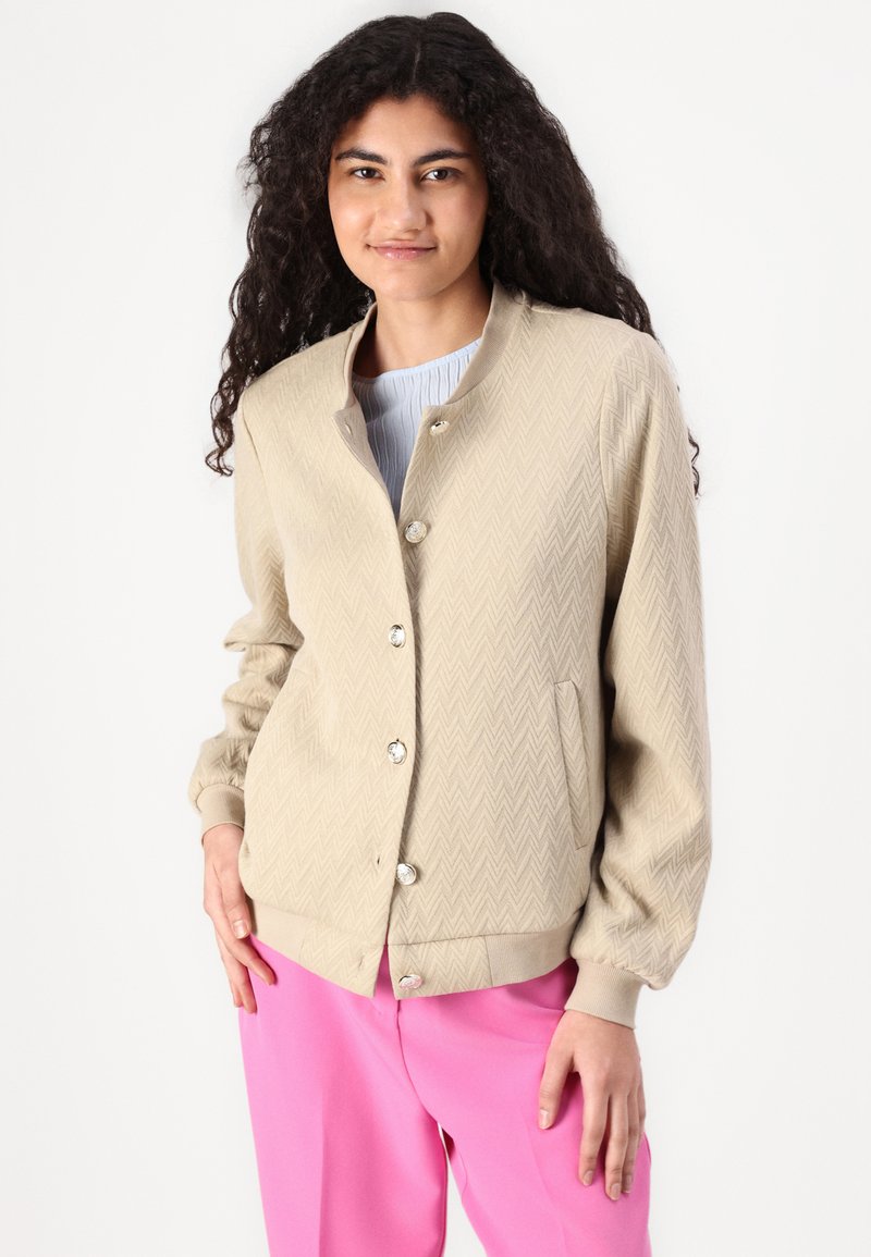 Giacca bomber beige con motivo a spina di pesce testurizzato, chiusura con bottoni e polsini a coste. Indossata sopra un top azzurro chiaro e pantaloni rosa.