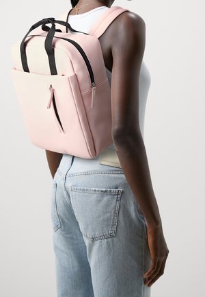 Femme portant un sac à dos rose clair avec des détails de fermeture éclair noirs, un haut sans manches blanc et un jean en denim bleu clair, vue de dos.