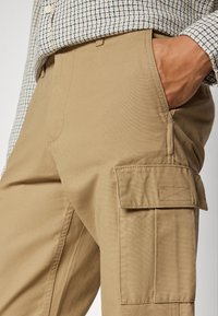 Khaki cargo nohavice s úzkym strihom, s bočnými vreckami, prešívaním a hladkým povrchom. V kombinácii s kockovanou košeľou.