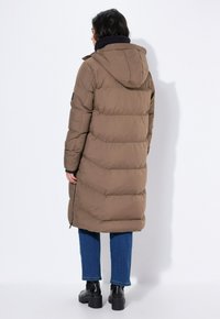 Manteau en duvet marron avec capuche, présentant des sections matelassées horizontales et une coupe longueur genoux. Porté par-dessus un jean bleu et des bottines noires.