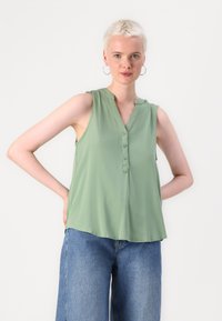 Blusa sem mangas verde-claro com decote em V, fecho de botão e tecido suave. Combinada com jeans de perna larga azul-claro. Design simples.