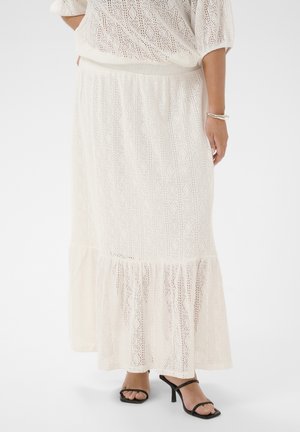 Femme portant une jupe longue en dentelle crème et un haut assorti, accompagnés de sandales noires à talons hauts avec brides et d'un bracelet argenté.