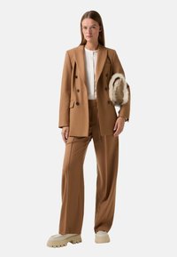 Bruine op maat gemaakte blazer met dubbele knopen, gecombineerd met bijpassende broek. Witte top eronder en beige schoeisel met een dikke zool.