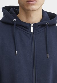 Navy zip-up hoodie with a drawstring hood, featuring a textured fabric, silver hardware, and flatlock stitching along the seams.