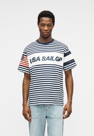 Mladý muž oblečený v námornícky modrom a bielom pruhovanom tričku "USA SAIL GP" s americkou vlajkou na ľavom rukáve a svetlomodrých džínsoch.