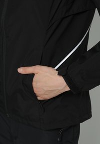 Mano in tasca della giacca nera con striscia riflettente e polsino in Velcro, zip parzialmente visibile su giacca e pantaloni.