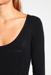 Musta pitkähihainen toppi, jossa on syvä avara kaula-aukko ja teksturoitu yksityiskohta. Topissa on hienovarainen kiiltävä kuvio ja joustava neulekangas.