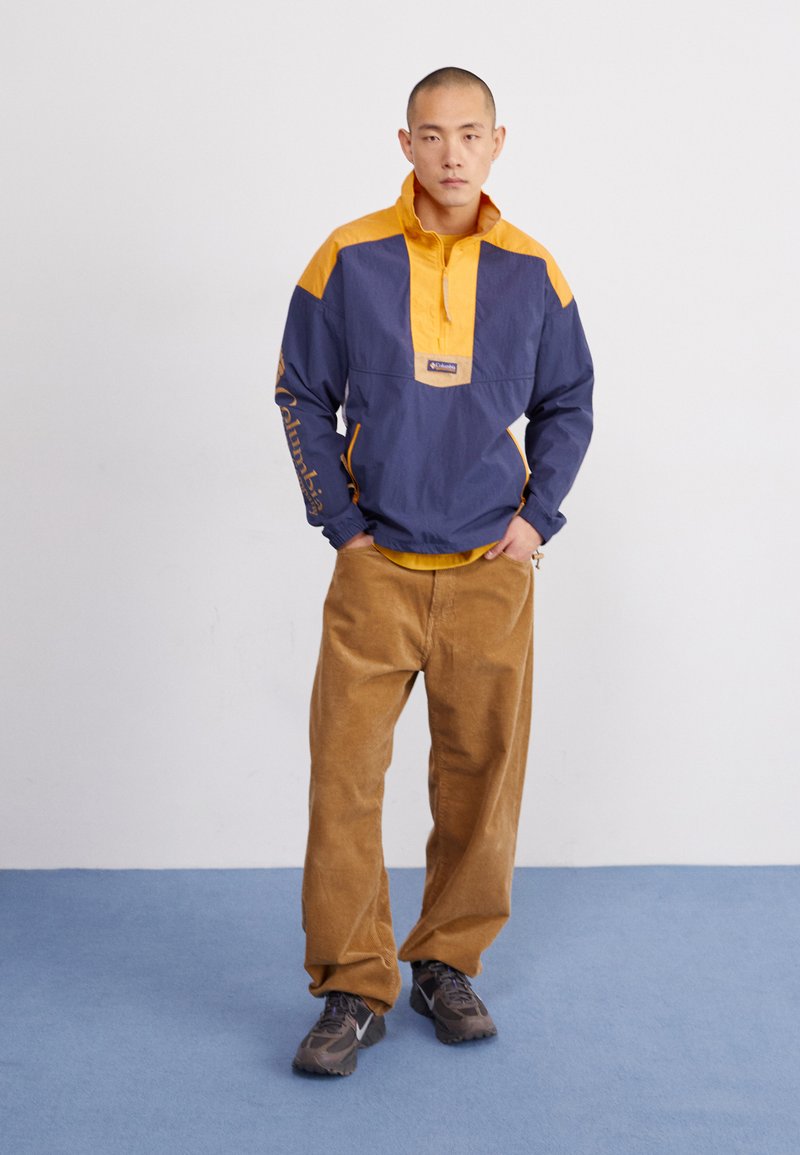 Kwart-ritsjack in kleurblokken, marineblauw en geel, met ritszakken. Gepaard met een bruine corduroy broek en grijze sneakers op een blauwe achtergrond.