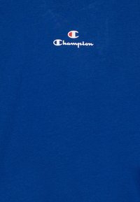 Camiseta de algodón azul con un pequeño logo de Champion en blanco y rojo centrado en el pecho. Textura suave y diseño de corte estándar.
