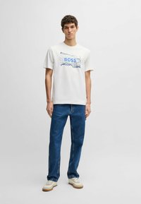 Hvid bomulds t-shirt med et blåt "BOSS" logo og bølgedesign, parret med højtaljede blå jeans og cremefarvede sneakers.