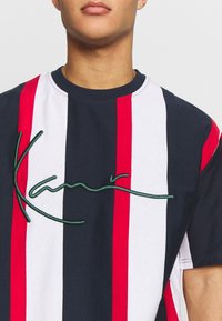 Tričko s krátkým rukávem s námořnickými, bílými a červenými vertikálními pruhy, se zeleným vyšitým textem přes hrudník. Klasický kulatý výstřih.