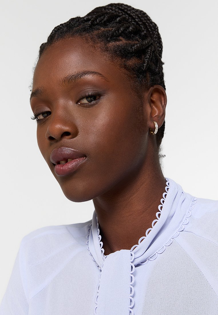 Blusa de color púrpura claro con cuello con volantes y detalle de lazo. Tela suave con una textura sutil, usada por un modelo con cabello trenzado.