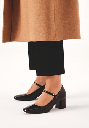 Chaussures à talons carrés en cuir verni noir avec un bout arrondi et une bride à la cheville ornée d'une petite boucle dorée. Portées avec un pantalon noir.