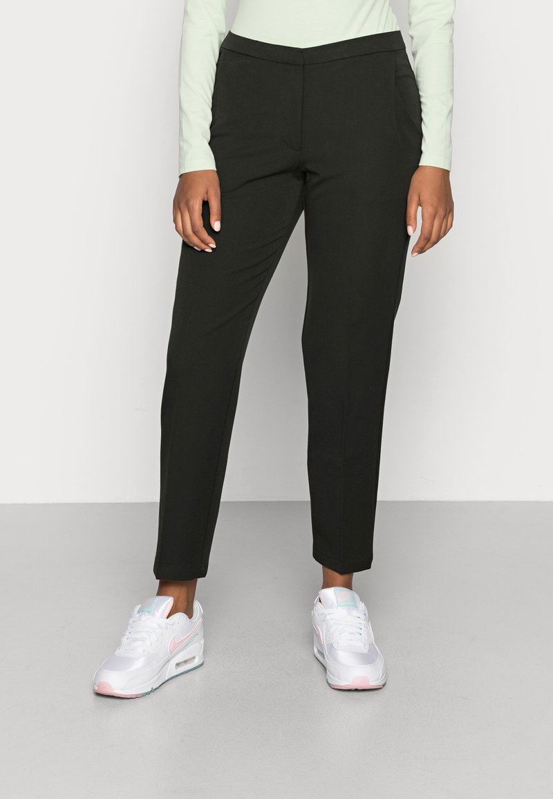 Zwarte op maat gemaakte broek met een slanke pasvorm, gladde stof en zijzakken, gecombineerd met witte sneakers met roze accenten.