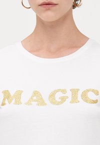 Hvit T-skjorte med korte ermer, med ordet "MAGIC" i store gullglitrende bokstaver over brystet. Mykt bomullsstoff.