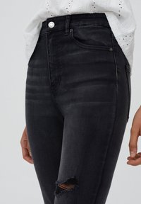 Mustat farkut, joissa on kapea istuvuus, kulutettu polvivyöleikkaus, tikkauksilla koristeltu yksityiskohta ja hopeinen nappi vyötäröllä.