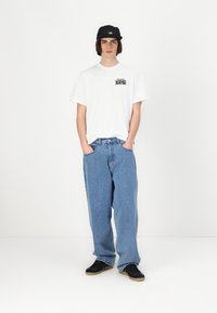 Hvid t-shirt med sort grafik, løse blå denimjeans, sort kasket og sorte sneakers med gummisål; model står med hænderne i lommerne.