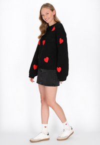 Schwarzer Strickpullover mit roten Herzmustern, kombiniert mit einem schwarzen Jeanrock und weißen Sneakers. Rote Herzakzente auf den Kniestrümpfen.