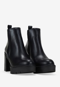Botas de tobillo de plataforma de cuero negro con tacones de bloque, paneles laterales elásticos y tiradores en un fondo blanco.