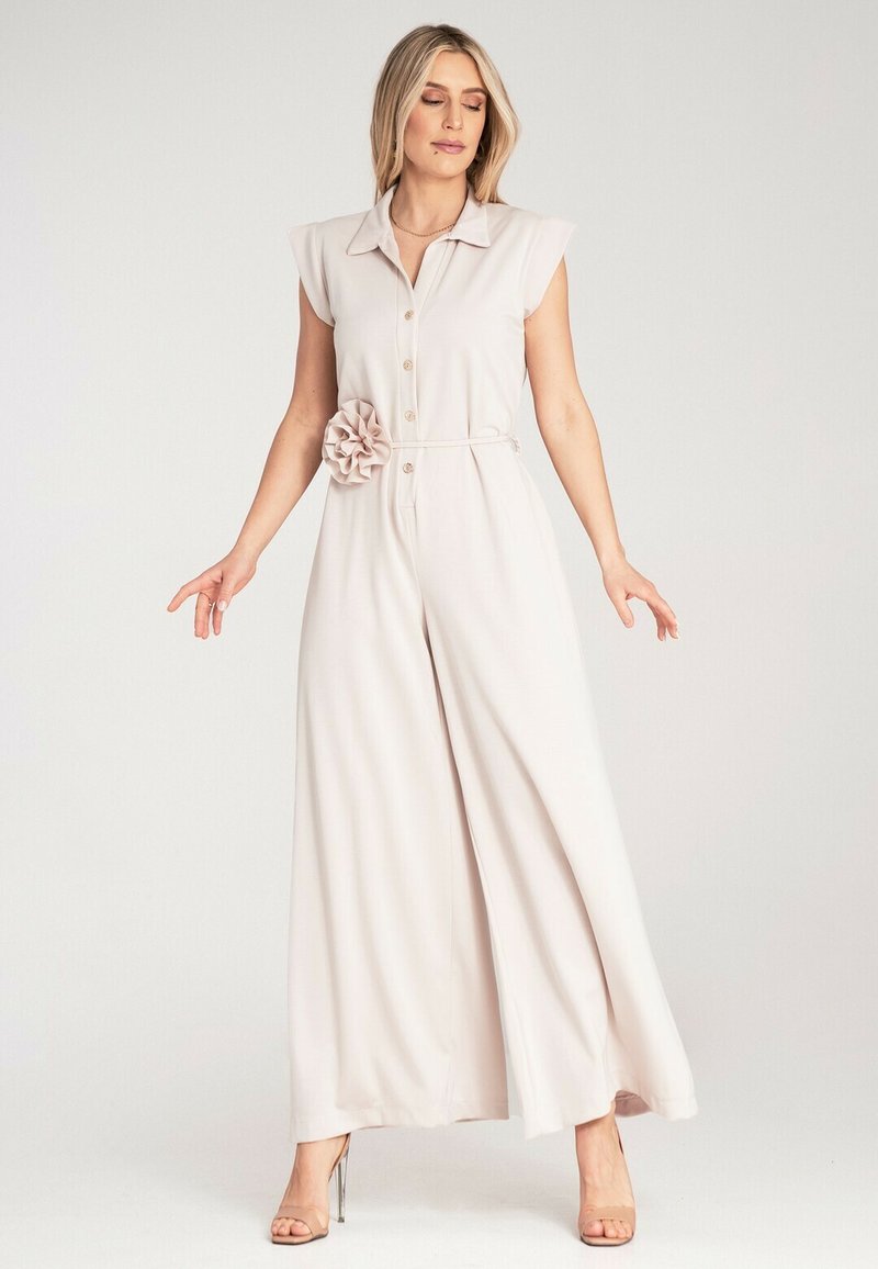 Jasnoróżowy kombinezon z krótkimi rękawami, zapięciem na guziki, materiałowym paskiem i dekoracyjnym akcentem w postaci kwiatu w talii. Luźny, szeroki krój nogawek.