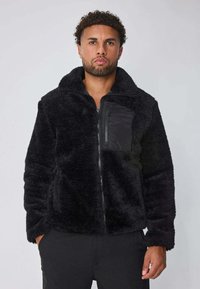 Homme portant une veste en polaire noire avec une poche poitrine zippée et les mains dans les poches latérales, se tenant devant un fond clair uni.