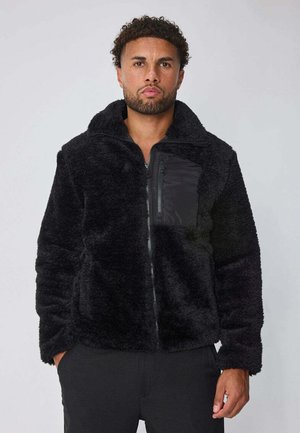 Homme portant une veste en polaire noire avec une poche poitrine zippée et les mains dans les poches latérales, se tenant devant un fond clair uni.