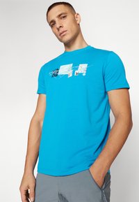 Camiseta de color azul brillante, hecha de una tela suave, que presenta un diseño gráfico con varias formas e imágenes en tonos blancos y azul claro en la parte frontal.