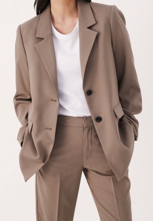 Person trägt einen taupefarbenen Anzug mit Blazer und passenden Hosen über einem weißen Rundhals-T-Shirt, Hände in den Taschen, vor einem schlichten hellen Hintergrund.