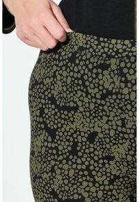 Una mano che regola la vita di pantaloni neri con un motivo a pois irregolari verdi, indossando una maglietta nera con cuciture gialle sulle maniche.